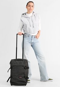 Musta kangaspintainen matkalaukku, jossa on useita hihnoja ja logo, seuranaan henkilö harmaassa hupparissa ja vaaleansinisissä farkuissa.