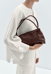 Brown leather handbag with soft texture and a structured design, featuring dual handles, held against a light-colored linen top.