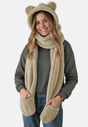 Beige fleece sjal med hætte, der har bjørneører, lommer i enderne og en blød, tekstureret overflade. Båret over en mørkegrå bluse.