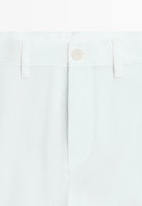 Hvid buksekant med knaplukning, bæltestropper og dækket lynlås.