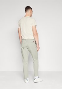 Jasno beżowa koszulka, widok z tyłu, zestawiona z jasnoszarymi spodniami i białymi tenisówkami z zielonymi akcentami. Miękka tkanina, casualowy design.