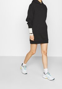 Schwarzes Hoodie-Kleid mit einer Kängurutasche, langen Ärmeln und Logo-Print entlang des Arms. Dazu passende weiße Sneakers mit bunten Akzenten.