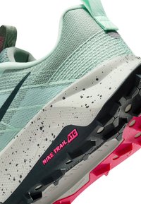 Jasnozielone buty do biegania w terenie Nike z nakrapianą białą podeszwą środkową, czarną i różową chropowatą podeszwą zewnętrzną oraz etykietą "NIKE TRAIL ATG" z boku.