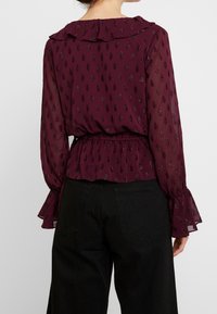 Blusa bordeaux con un colletto arricciato, vita elastica e maniche lunghe con polsini svasati. Tessuto trasparente con un design a motivo sottile.