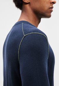 Tumši zils džemperis ar apaļu apkakli, ribota tekstūra, piegulošs siluets un kontrastējošas zaļas šuves gar plecu šuvēm.