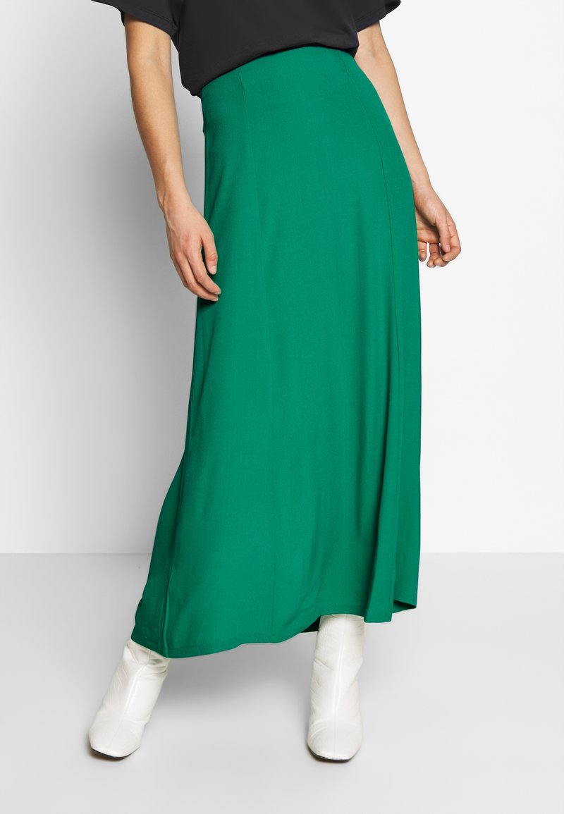 Groene maxirok van gladde stof, met een hoge taille en een rechte snit. Gecombineerd met witte enkellaarsjes voor contrast.