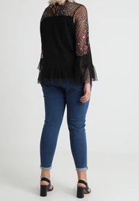 Haut noir avec broderie florale et manches en polka dots transparentes, associé à un jean bleu avec manches roulées et des sandales noires à talons hauts.