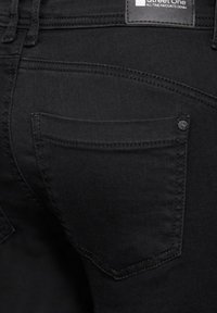 Svart denim jeans med en slät yta, med en bakficka och synliga sömnadsdetaljer. Inkluderar en märkesetikett i tyg vid midjan.