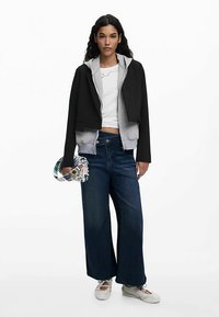 Gekürzter schwarzer und grauer Hoodie, weißes Shirt und weite dunkelblaue Jeans aus Denim. Zubehör umfasst eine bunt gemusterte Tasche und helle Schuhe.