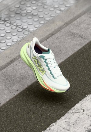 Weißer und grüner Laufschuh mit gelber Sohle und "HOKA"-Logo, schwebt über Asphalt in der Nähe eines Bürgersteigrands und einer strukturierten taktilen Leitlinie.