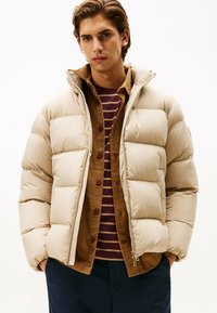 Beżowa kurtka puchowa z wysokim kołnierzem i pikowanym wzorem, nałożona na brązową koszulę z sztruksu i bordową bluzkę w paski, na białym tle.
