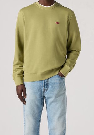 Homme portant un sweat-shirt Levi's vert olive et un jean bleu clair, la main dans la poche, debout devant un fond uni.