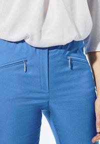 Pantalon bleu en tissu lisse avec deux poches zippées et matériel métallique. La taille est plate avec une finition soignée.