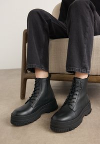 Bottines en simili cuir noir avec une semelle épaisse et texturée, un design à lacets et une fermeture éclair sur le côté pour un accès facile.