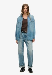 Jasnoniebieska kurtka dżinsowa z dwoma kieszeniami na klatce piersiowej, brązowy top i luźne jasnoniebieskie dżinsy, zestawione z czarnymi butami. Gładka faktura.