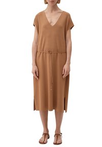 Vrouw in een camel-kleurige jurk tot de knie met korte mouwen, V-hals, dunne tailleband, zijsplits en bruine platte sandalen.
