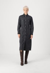 Tmavodžínová košeľová šaty s dlhými rukávmi, predným gombíkovým zapínaním, dvoma náprsními vreckami a dĺžkou po stred lýtok, skombinované s hnedými vysokými čižmami.