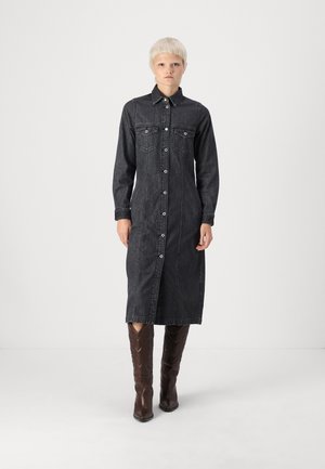 Abito camicia in denim scuro con maniche lunghe, chiusura frontale con bottoni, due tasche sul petto e lunghezza a metà polpaccio, abbinato a stivali marroni al ginocchio.