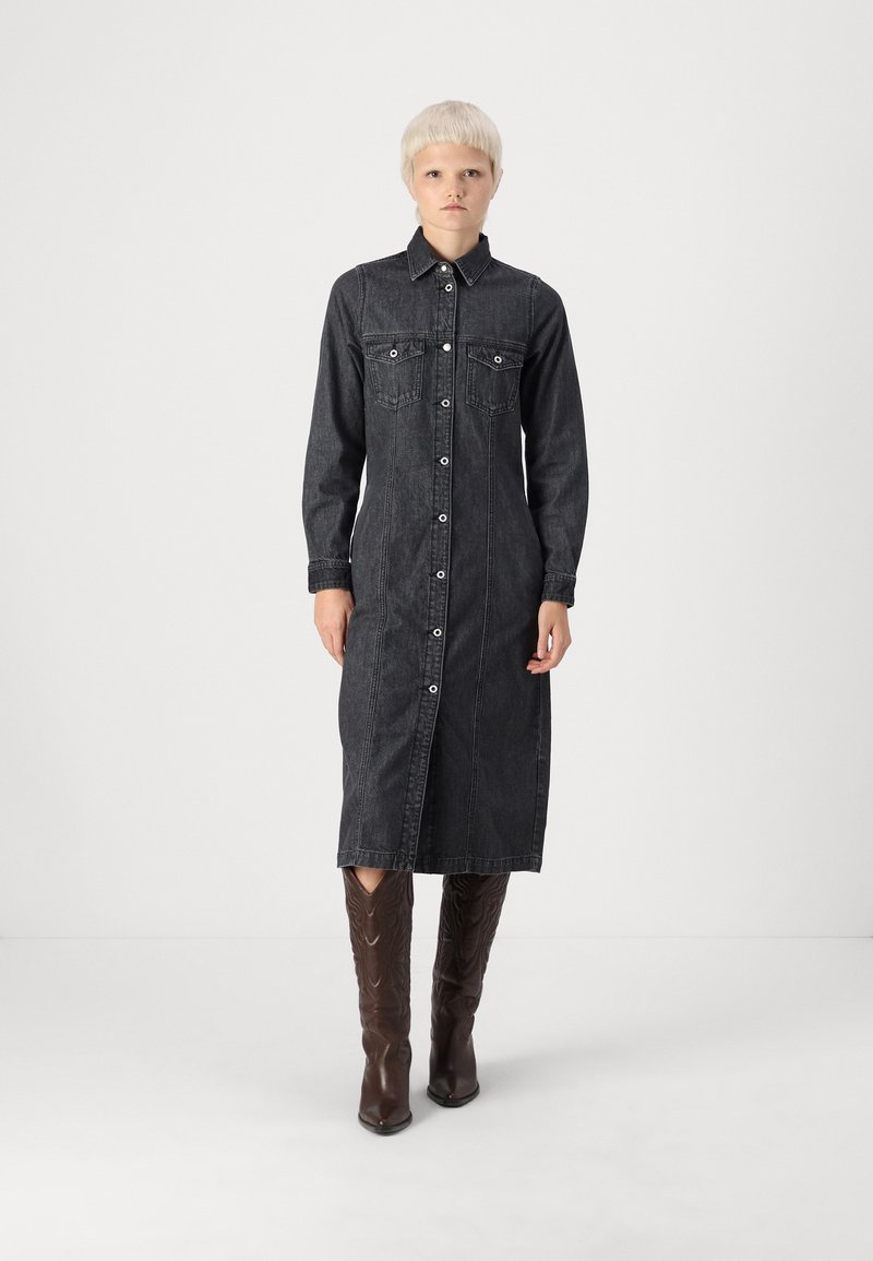 Tmavodžínová košeľová šaty s dlhými rukávmi, predným gombíkovým zapínaním, dvoma náprsními vreckami a dĺžkou po stred lýtok, skombinované s hnedými vysokými čižmami.