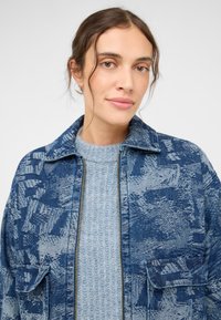 Veste en denim bleu avec des motifs abstraits, fermeture éclair et deux poches avant sur un pull à rayures bleu clair. Texture de tissu douce.