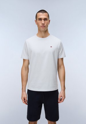 Ung mand stående iført en simpel hvid kortærmet T-shirt med lille rødt logo på brystet og sorte shorts, neutral lys baggrund.