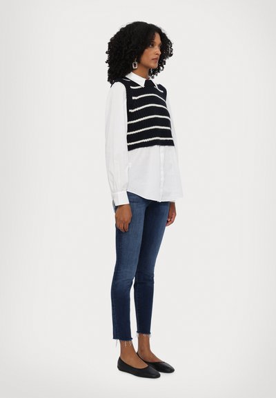 Fekete-fehér csíkos kötött cropped top egy fehér ing felett, sötét farmer skinny nadrággal és fekete lapos cipővel párosítva.