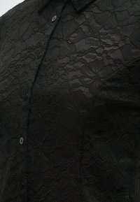 Țesătură din dantelă neagră cu model floral suprapus peste un design de cămașă cu guler și nasturi, într-o secțiune apropiată a articolului de îmbrăcăminte.