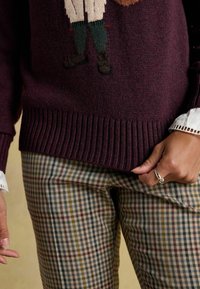 Burgunderfarbener Strickpullover mit geripptem Saum und gestickter Figur; getragen mit beigen und karierter Hose, die grüne, gelbe und rote Streifen aufweist.