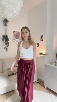 Bílý krátký top bez rukávů v kombinaci s dlouhou vínově červenou plisovanou sukní. V pozadí se nachází světlá pohovka a dekorativní prvky.