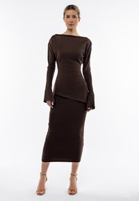 Abito in maglia a coste marrone con maniche lunghe, scollo a barca, orlo asimmetrico e silhouette aderente. Abbinato a sandali nude con cinturini.