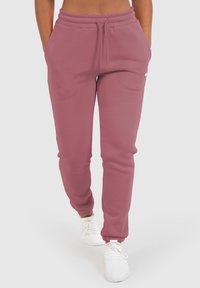 Rosa Jogginghose mit elastischem Bund, Seitentaschen und gerippten Säumen, aus weichem Stoff. Getragen mit weißen Sportschuhen.