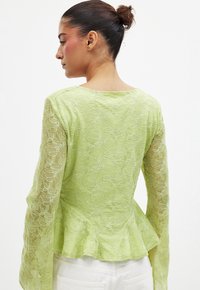 Hellgrünes Peplum-Oberteil mit langen Spitzenärmeln, die mit floralen Stickereien verziert sind, und einem runden Ausschnitt; durchscheinender strukturierten Stoff.
