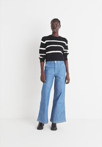 Zwart-wit gestreepte trui, blauwe wijde jeans, gedragen met zwarte schoenen. Minimalistisch ontwerp tegen een effen witte achtergrond.