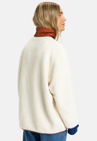 Fleece pullover in crèmekleur met een gestructureerde afwerking, voorzien van een bruine kraag en blauwe mouwaccenten. Losse, boxy fit, heuplengte ontwerp.