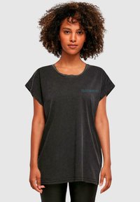Schwarzes, oversized T-Shirt mit rundem Halsausschnitt, umgeschlagenen kurzen Ärmeln und einer verblassten Textur. Mit türkisfarbenem Text, der "Iron Maiden" liest.