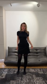 Woman with blonde hair wearing a black dress and tights standing in a minimalist living room with a dark gray sofa and patterned rug.