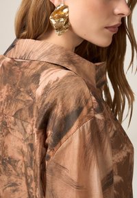 Camisa marrón con estampado, de textura sedosa y con cuello notch; presenta un acento dorado y texturizado en forma de pendiente en la oreja del modelo.