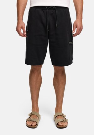 Uomo con pantaloncini neri al ginocchio con tasche con zip e sandali beige con fibbia, in piedi contro uno sfondo semplice.