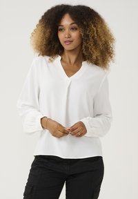 Blouse blanche, tissu texturé, décolleté en V, manches longues avec poignets froncés, ornée de détails de boutons subtils, associée à un pantalon sombre.