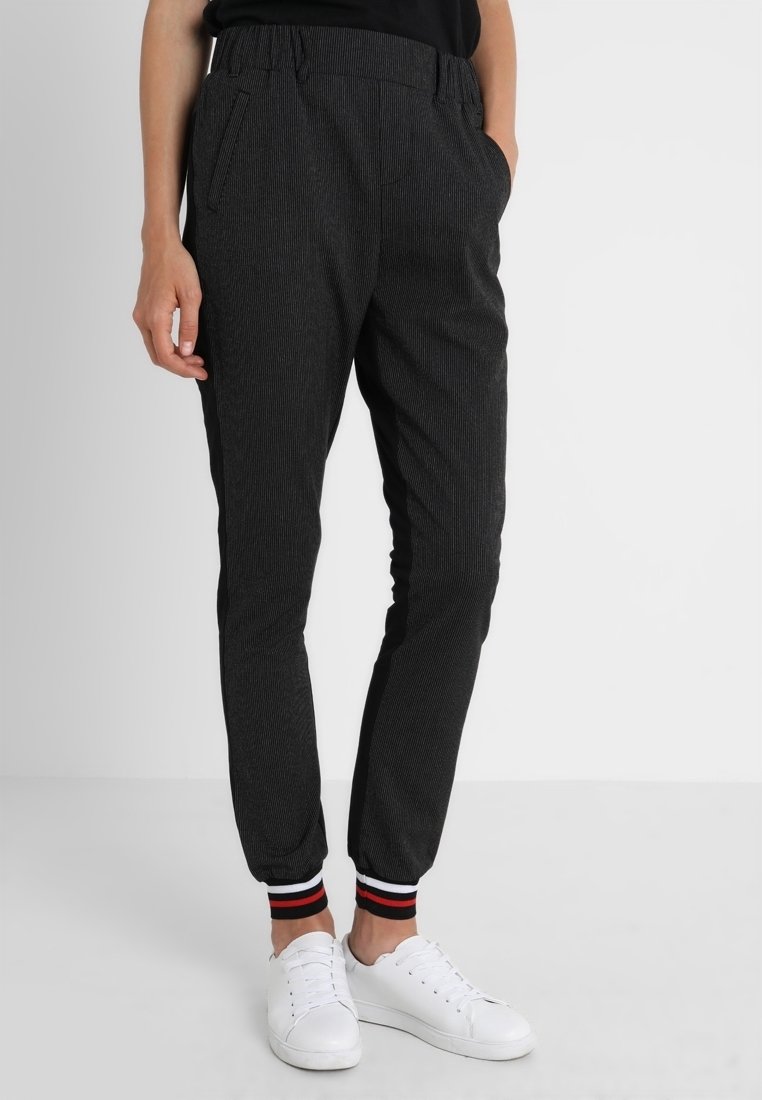 Moitié inférieure d'une personne portant un pantalon de jogging foncé fuselé avec des poignets élastiques montrant des rayures noires, blanches et rouges, associé à des baskets blanches.