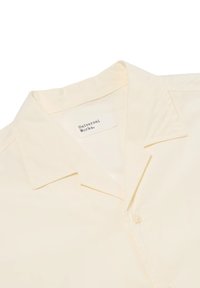 Camicia a maniche corte giallo chiaro con colletto, dotata di un bottone, morbida al tatto e con un'etichetta che dice "Universal Works."