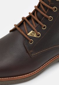 Botas de cuero marrón con un acabado texturizado; presenta cordones marrones, ojales de latón y un contraste de costura blanca a lo largo de la suela.