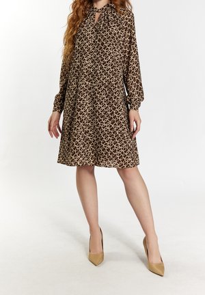 Kobieta w brązowej, wzorzystej sukience do kolan z długimi rękawami i beżowych szpilkach, stojąca na białym tle.