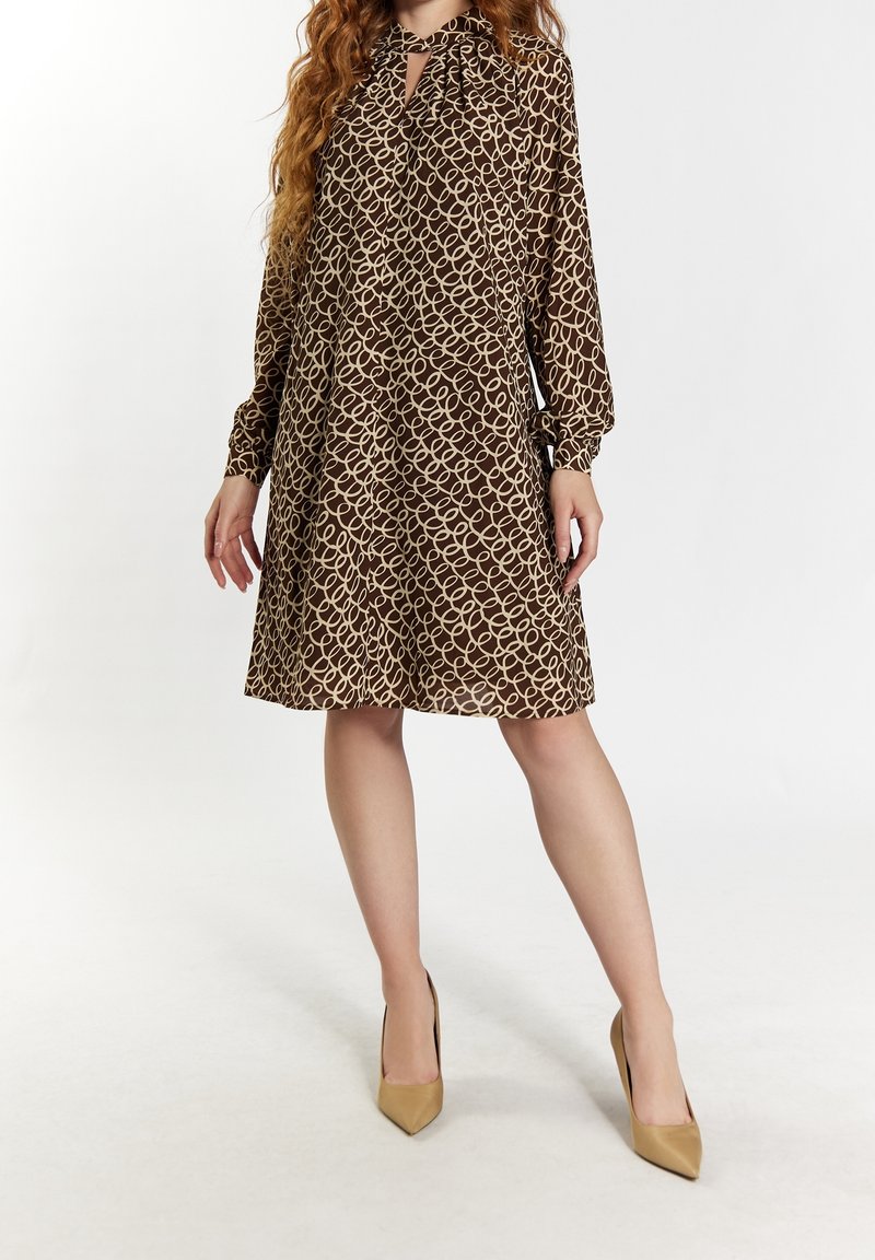 Femme portant une robe marron à motifs longueur genoux avec manches longues et des talons hauts beige, debout devant un fond blanc.