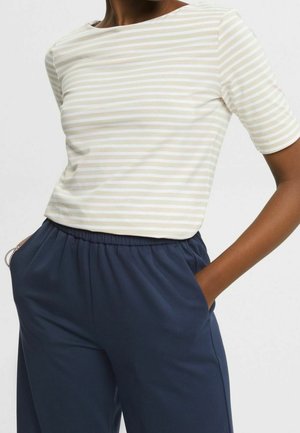 Persoon die een beige-wit gestreept T-shirt met korte mouwen draagt, ingestopt in marineblauwe broek met elastische taille, handen in de zakken.