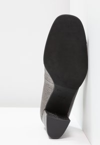 Chaussure à talon grise présentant une tige texturée et une semelle lisse avec une base en caoutchouc noir. Le talon est de forme carrée et légèrement effilé.