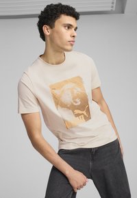 T-shirt beige con una grande grafica arancione di un gatto ruggente; scollatura rotonda; maniche corte; il tessuto appare morbido e liscio; indossato con jeans scuri.