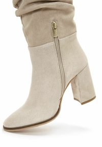 Stivale beige in scamosciato alto fino al ginocchio, con punta affusolata, tacco a blocco spesso e dettaglio della zip esterna. Texture morbida e design aderente.