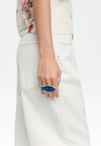 Blauwe hars ring in de vorm van een vis met textuurdetails en gouden accenten, gedragen op de wijsvinger, tegen een achtergrond van witte broeken.