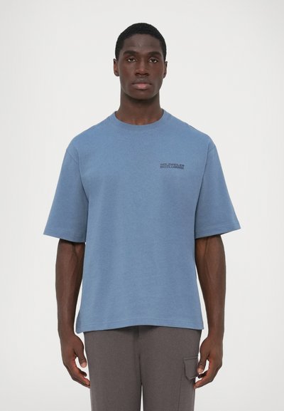 T-shirt bleue en coton avec des manches courtes, encolure ronde, et un logo imprimé en police foncée sur le côté gauche. Porté avec un pantalon gris ample.